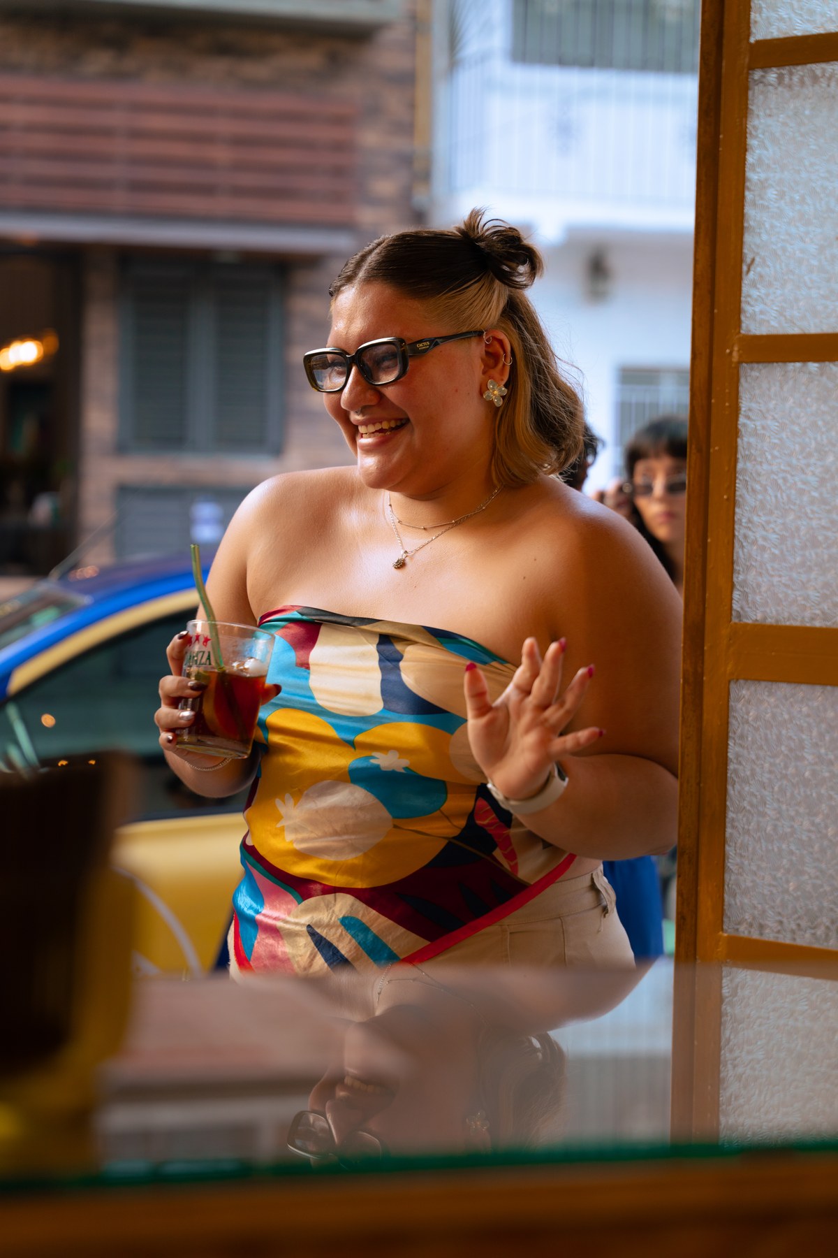 Patron smiling and gesturing through the cafe doorway with branded drink in hand and glass reflection in foreground